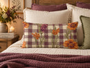 A rectangular decorative throw pillow with a purple and beige gingham pattern and orange floral print, styled on a bed with other pillows and a blanket.