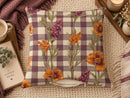 A square throw pillow with a purple and cream gingham pattern overlaid with orange and purple wildflower illustrations, styled on a beige knit blanket with a mug, book, and candle nearby.