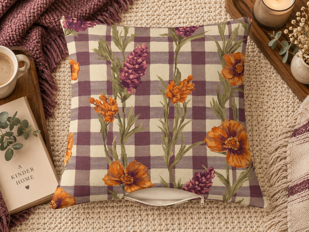 A square throw pillow with a purple and cream gingham pattern overlaid with orange and purple wildflower illustrations, styled on a beige knit blanket with a mug, book, and candle nearby.