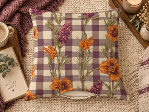 A square throw pillow with a purple and cream gingham pattern overlaid with orange and purple wildflower illustrations, styled on a beige knit blanket with a mug, book, and candle nearby.