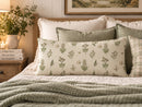 A decorative lumbar pillow with a green botanical and bird pattern on a cream background, styled on a bed with other neutral-toned pillows and a textured green blanket in a cozy bedroom setting.