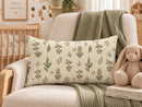 A rectangular throw pillow with a botanical leaf pattern on a cream background, styled on a beige armchair in a nursery setting next to a plush bunny and a green blanket.