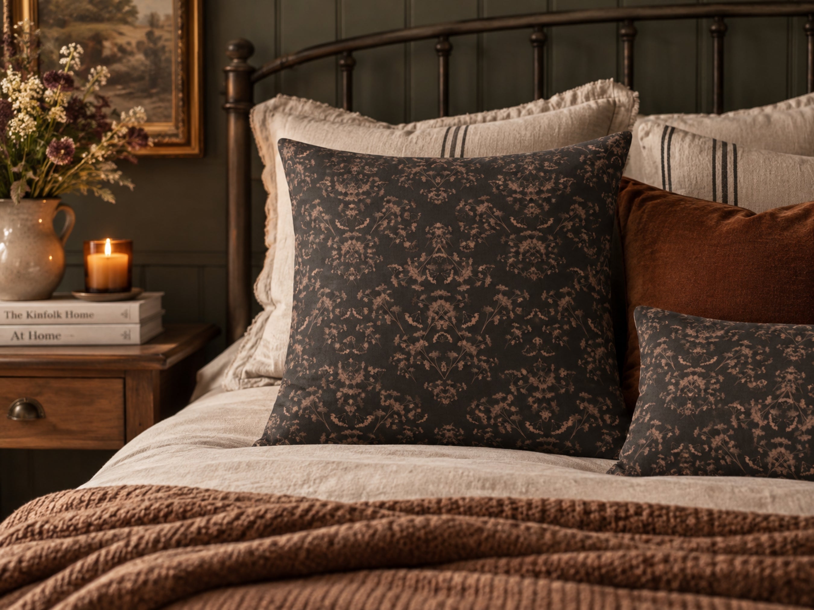 A styled bedroom scene with a dark charcoal damask-patterned pillow as the main product, layered with cream and rust accent pillows on a bed with a brown knit blanket. A wooden nightstand with a candle and books is visible on the left.