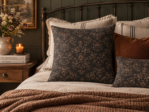 A styled bedroom scene with a dark charcoal damask-patterned pillow as the main product, layered with cream and rust accent pillows on a bed with a brown knit blanket. A wooden nightstand with a candle and books is visible on the left.