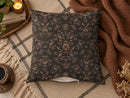A square throw pillow with a dark charcoal gray background and a warm brown damask floral pattern, styled on a beige plaid rug with a brown knit blanket, candles, and dried flowers.