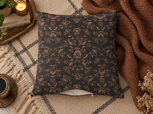 A square throw pillow with a dark charcoal gray background and a warm brown damask floral pattern, styled on a beige plaid rug with a brown knit blanket, candles, and dried flowers.