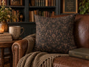 A square throw pillow with a dark brown and beige damask pattern, styled on a brown leather armchair in a cozy, book-filled library setting.