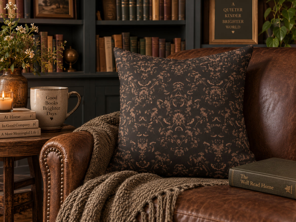 A square throw pillow with a dark brown and beige damask pattern, styled on a brown leather armchair in a cozy, book-filled library setting.