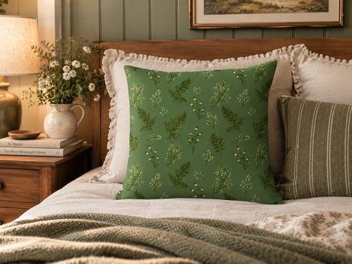A green decorative throw pillow with a delicate botanical leaf pattern, styled on a bed with white and striped pillows in a warm, cozy bedroom setting.
