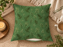A square throw pillow with a green botanical fern pattern, styled on a beige woven rug with a candle, mug, and blanket in the background.