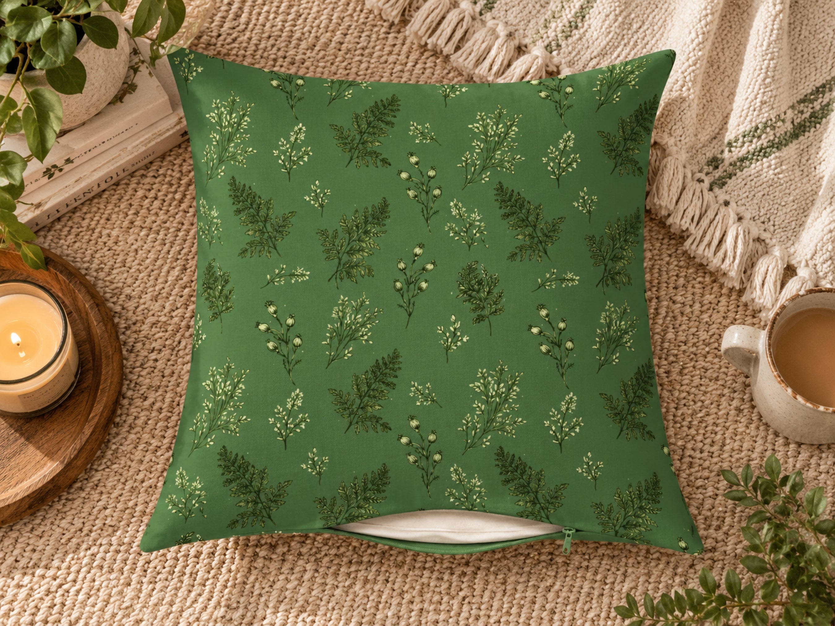 A square throw pillow with a green botanical fern pattern, styled on a beige woven rug with a candle, mug, and blanket in the background.