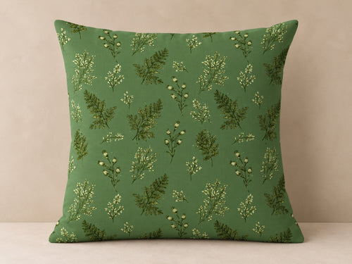 A square throw pillow with a green background and a repeating pattern of botanical ferns and small white flowers, set against a neutral beige background.