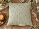A square throw pillow with a sage green botanical pattern on a pale mint background, styled on a woven jute rug with a candle, books, and a throw blanket.