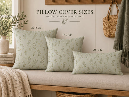Three decorative throw pillows with a light sage green and beige botanical leaf pattern are arranged on a beige upholstered wooden bench. The scene includes size callouts reading 22x22, 18x18, and 20x12, along with home decor props like a woven basket bag, a throw blanket, and a plant in a vase.