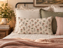 A cozy bedroom scene with a floral throw pillow centered on a bed, surrounded by cream pillows, a green accent pillow, a pink striped pillow, a quilted blanket, and a bedside table with flowers and books.