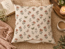 A square throw pillow with a vintage-style floral and butterfly print on a cream background, styled on a woven jute mat with a knit blanket, an open book, a candle, and a cup of coffee.