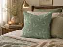 A square decorative throw pillow with a sage green butterfly pattern, styled on a bed in a warm, cozy bedroom setting.