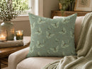 A square throw pillow with a sage green and gray butterfly pattern, styled on a beige armchair in a cozy living room setting with a throw blanket, candles, and a vase.