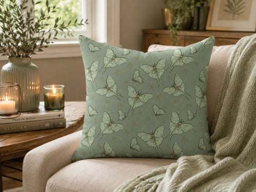 A square throw pillow with a sage green and gray butterfly pattern, styled on a beige armchair in a cozy living room setting with a throw blanket, candles, and a vase.